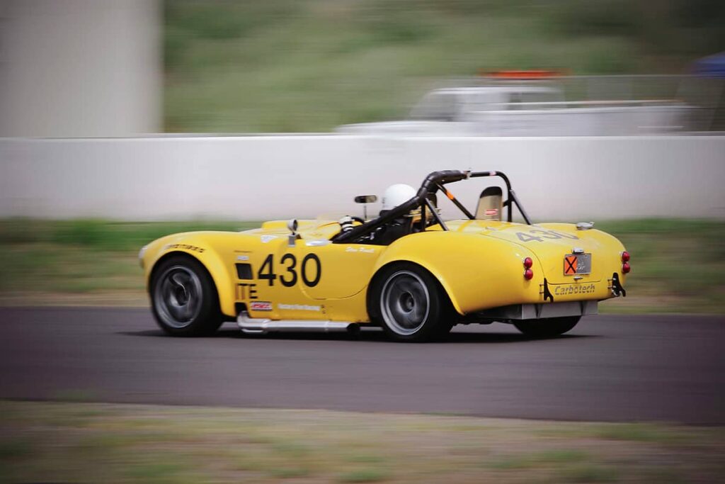 Yellow AC Cobra open top roadster with a roll cage at the local race track